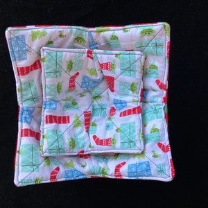 May include: Two square pot holders with a white background and a pattern of green Christmas trees, red and white stockings, and green and blue gift boxes. The pattern also includes small green aliens with big eyes. The pot holders are quilted and have a red border.