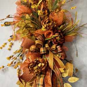 May include: A fall-themed floral arrangement featuring a mix of faux flowers, leaves, and ribbons in shades of orange, gold, and brown. The arrangement includes decorative bows and accents, creating a festive and seasonal display.