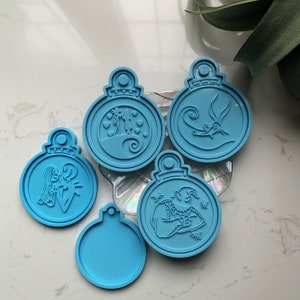 Puede incluir: Cinco moldes de adornos navideños de color azul claro con varios diseños. Un molde está en blanco, mientras que los otros presentan personajes y escenas de una popular película de animación. Los moldes son redondos con un lazo en la parte superior para colgar.