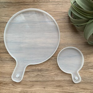 May include: Two clear silicone moulds for resin casting. The larger mould is shaped like a round tray with a handle. The smaller mould is a smaller version of the larger mould.