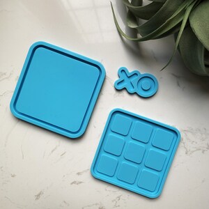 May include: Three blue silicone molds for resin crafts. One mold is a square with a smooth surface, another is a square with nine square indentations, and the last is a small mold shaped like the letters "XO".