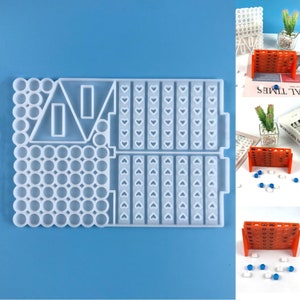 May include: A clear, rectangular silicone mold for crafting. The mold features various shapes, including circles, triangles, rectangles, and heart-shaped columns. The mold is designed for creating game pieces and decorative elements. The background is a solid light blue.