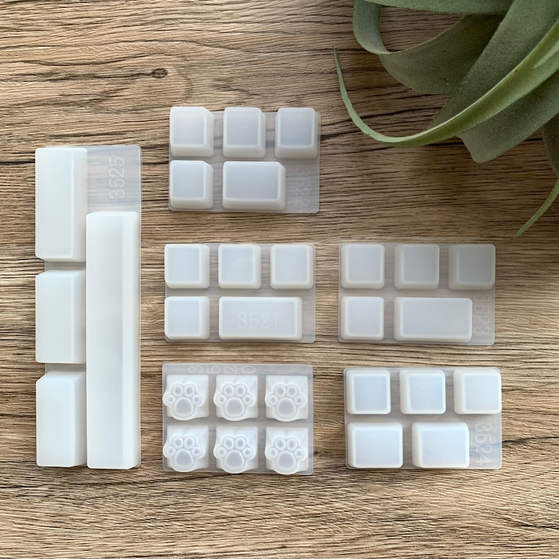May include: A collection of clear silicone molds for crafting custom keyboard keycaps. The molds feature various shapes, including rectangular, square, and paw print designs. The molds are arranged on a wooden surface.