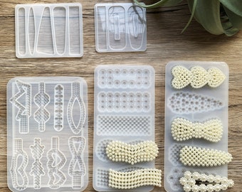 Könnte beinhalten: Sechs transparente Silikonformen zum Herstellen von Haarklammern. Die Formen sind in zwei Reihen mit jeweils drei Formen angeordnet. Die obere Reihe zeigt drei verschiedene Formen von Haarklammern. Die untere Reihe zeigt drei verschiedene Designs von Haarklammern mit perlmuttartigen Details.