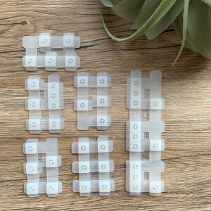 May include: A collection of translucent white silicone keypads with square buttons and cross-shaped markings. The keypads are arranged on a wooden surface, with a green plant in the background. Some keypads have the numbers 3523, 3524, and 3298 printed on them.