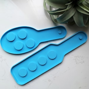 May include: Two bright blue silicone moulds. One is oval-shaped with five circular indentations, and the other is rectangular with four circular indentations. Both have a handle-like extension with a small circular indentation. The moulds are designed for crafting.