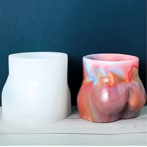 May include: A white silicone mould and a finished decorative pot. The pot is shaped like a torso with a rounded base and is painted with swirls of pink, orange, and blue. The mould is a solid white colour.