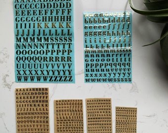 May include: Gold-coloured alphabet stickers in various sizes and fonts. The stickers are arranged on light blue and tan backing sheets. The letters include uppercase and lowercase, ideal for craft and scrapbooking projects.