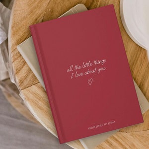 Op de afbeelding: Een bordeauxrood notitieboekje met de tekst "all the little things I love about you" en een klein hartje. Het notitieboekje heeft de tekst "FROM JAMES TO EMMA" onderaan.