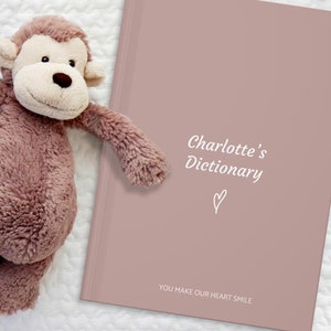 May include: A pink hardcover journal with the text "Charlotte's Dictionary" and a small heart symbol. The text "You make our heart smile" is printed at the bottom of the cover.