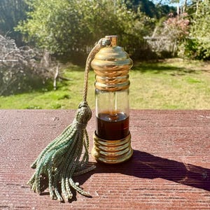 May include: A small, clear glass bottle with a gold cap and a green tassel. The bottle is filled with a dark brown liquid.