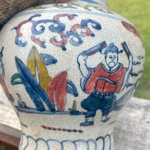 May include: A ceramic vase with a crackled glaze finish featuring hand-painted blue, red, and yellow illustrations. The design depicts a figure in traditional attire holding rectangular objects, surrounded by stylised foliage and cloud motifs.