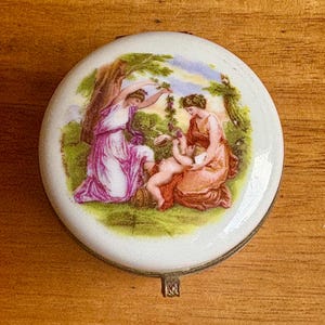 Könnte beinhalten: Eine runde Porzellan-Schmuckdose mit einem Scharnierdeckel. Der Deckel zeigt eine bemalte Szene mit drei Figuren in einem Garten. Die Figuren sind eine Frau in einem lila Kleid, eine Frau in einem rosa Kleid und ein Kind in einem weißen Kleid. Die Szene ist von grünem Laub und einem blauen Himmel umgeben.