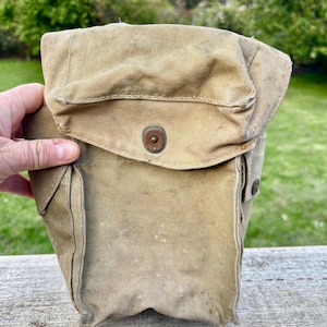 May include: A vintage tan canvas bag with a flap and metal snap closure. The bag is rectangular with a front pocket. The bag is held by a hand. The bag appears to be of military or utilitarian design.