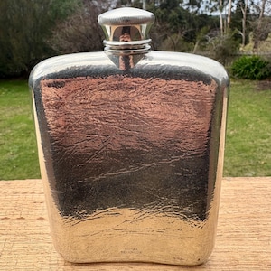 Puede incluir: Una petaca plateada con una superficie texturizada y un tapón de rosca. La petaca tiene bordes redondeados y forma rectangular. Las palabras "ROYAL SELANGOR" están impresas en la parte inferior.
