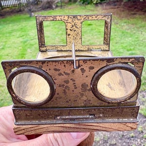 Può includere: Un visore vintage color ottone con due lenti rotonde e un'apertura rettangolare nella parte superiore. Il visore è in metallo e ha una base in legno.