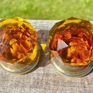 May include: Two amber-coloured, faceted glass door knobs with brass bases. The knobs have a geometric design and sit on a weathered wooden surface, with a blurred green background.
