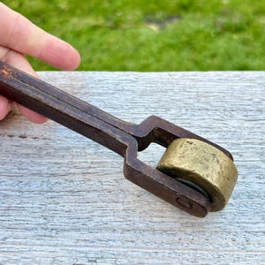 May include: An antique metal tool with a wooden handle and a brass roller. The tool is dark brown with a worn appearance, suggesting age and use. The roller is cylindrical and appears to be made of solid brass.