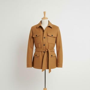 Puede incluir: Chaqueta de color marrón claro con cinturón en la cintura y cuatro bolsillos delanteros. La chaqueta tiene cuello y cierre de botones. La chaqueta se exhibe en un maniquí sobre un fondo blanco.