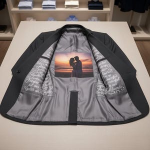 以下が含まれることがあります： 濃いグレーのスーツジャケット、グレーの裏地付き。 裏地には、夕焼け時のカップルの写真と手書きのテキストがプリントされています。 ジャケットは明るい色の表面に開いた状態で表示されています。