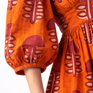 May include: Orange dress with a repeating pattern of maroon and cream-colored leaf-like shapes. The dress features puffed sleeves and a V-neckline. The fabric appears to be cotton or a similar material.