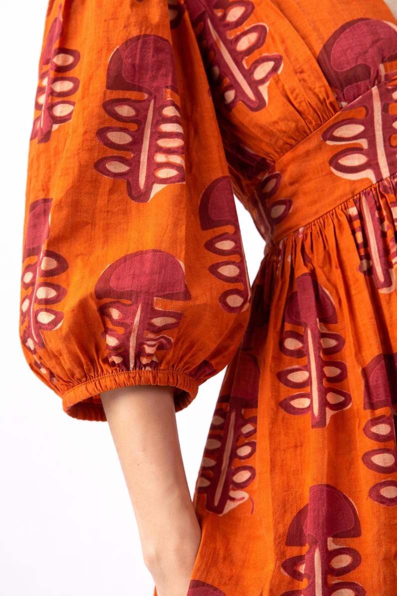 May include: Orange dress with a repeating pattern of maroon and cream-colored leaf-like shapes. The dress features puffed sleeves and a V-neckline. The fabric appears to be cotton or a similar material.