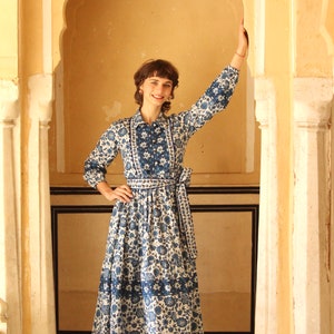 May include: A woman wearing a blue and white floral dress with a white collar and a blue sash. She is standing in a doorway with a yellow archway.