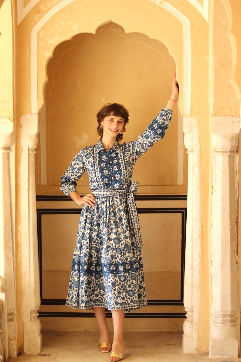 May include: A woman wearing a blue and white floral dress with a white collar and a blue sash. She is standing in a doorway with a yellow archway.