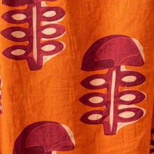 May include: Close-up of orange fabric with a repeating pattern of stylized, maroon-colored plant-like shapes. The shapes have a rounded top and a stem with oval-shaped details. The background is a solid orange color.