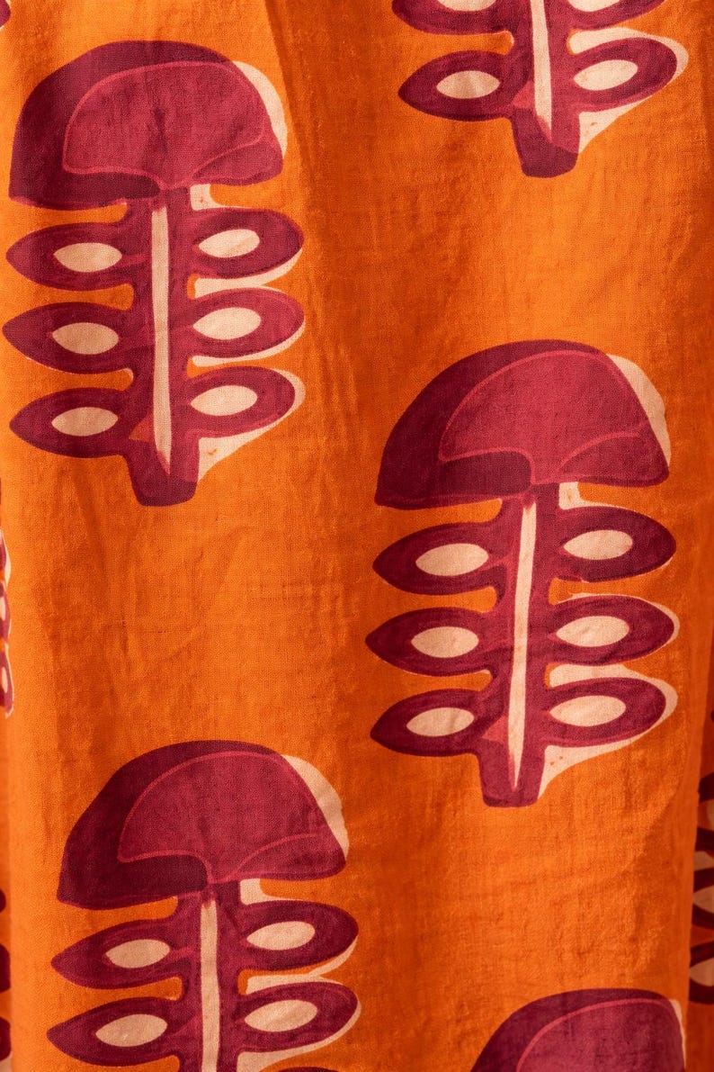 May include: Close-up of orange fabric with a repeating pattern of stylized, maroon-colored plant-like shapes. The shapes have a rounded top and a stem with oval-shaped details. The background is a solid orange color.