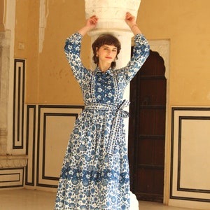 May include: A woman wearing a white and blue floral print dress with a long sleeve and a tie at the waist. The dress has a blue and white trim around the neckline and the hem. The woman is standing in front of a white pillar.