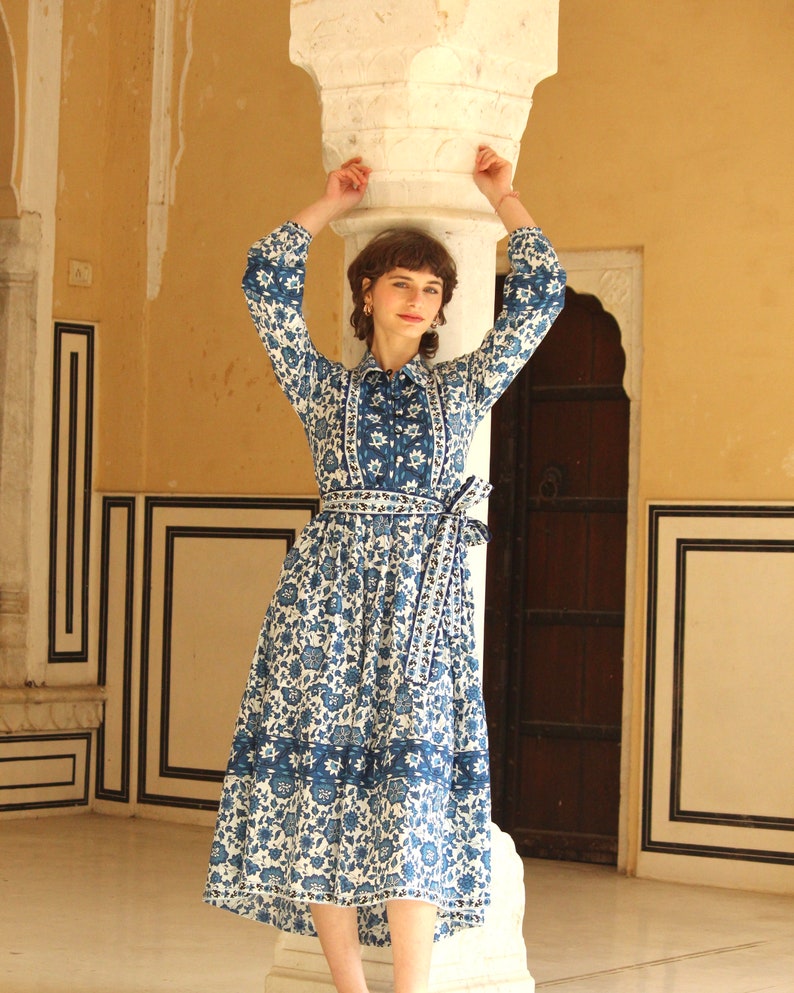 May include: A woman wearing a white and blue floral print dress with a long sleeve and a tie at the waist. The dress has a blue and white trim around the neckline and the hem. The woman is standing in front of a white pillar.