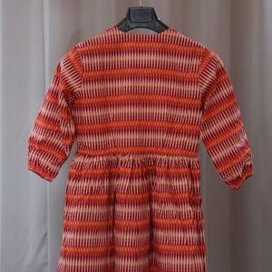 Op de afbeelding: Een rood-wit gepatroneerde jurk met lange mouwen en een geplooide rok. De jurk hangt aan een houten hanger.