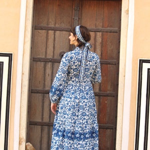 May include: A woman wearing a white dress with a blue floral pattern and a blue headband. The dress has a long sleeve and a gathered skirt. The woman is standing in front of a wooden door.