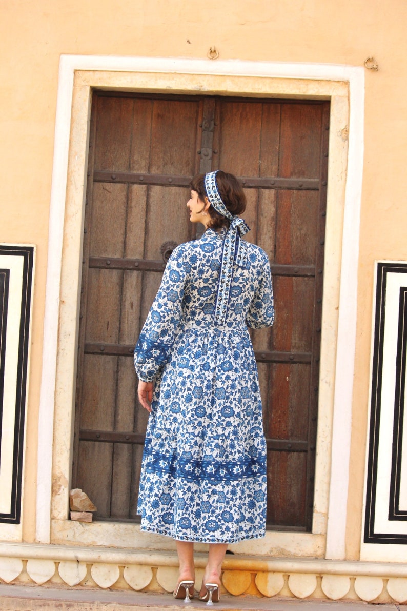 May include: A woman wearing a white dress with a blue floral pattern and a blue headband. The dress has a long sleeve and a gathered skirt. The woman is standing in front of a wooden door.