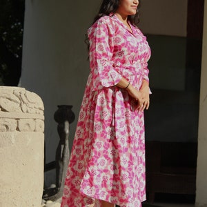 May include: A woman wearing a pink and white floral print dress with a long sleeve and a tie at the waist. She is standing on a wooden platform and wearing sandals.