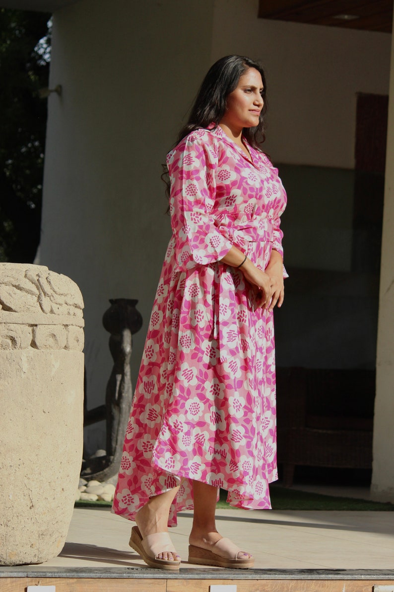 May include: A woman wearing a pink and white floral print dress with a long sleeve and a tie at the waist. She is standing on a wooden platform and wearing sandals.