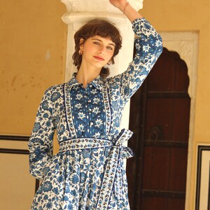 May include: A woman wearing a blue and white floral print dress with a long sleeve and a tie at the waist. The dress has a white background with blue floral patterns and a blue and white border.