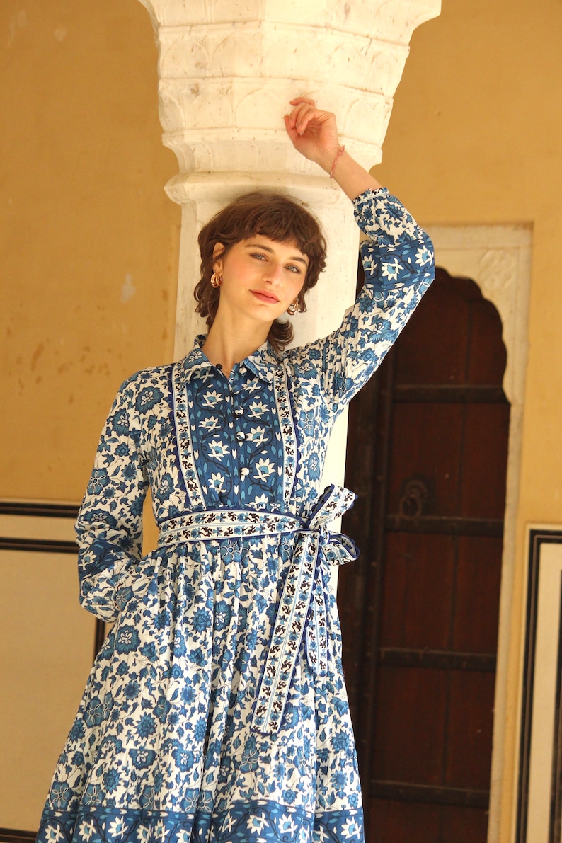May include: A woman wearing a blue and white floral print dress with a long sleeve and a tie at the waist. The dress has a white background with blue floral patterns and a blue and white border.