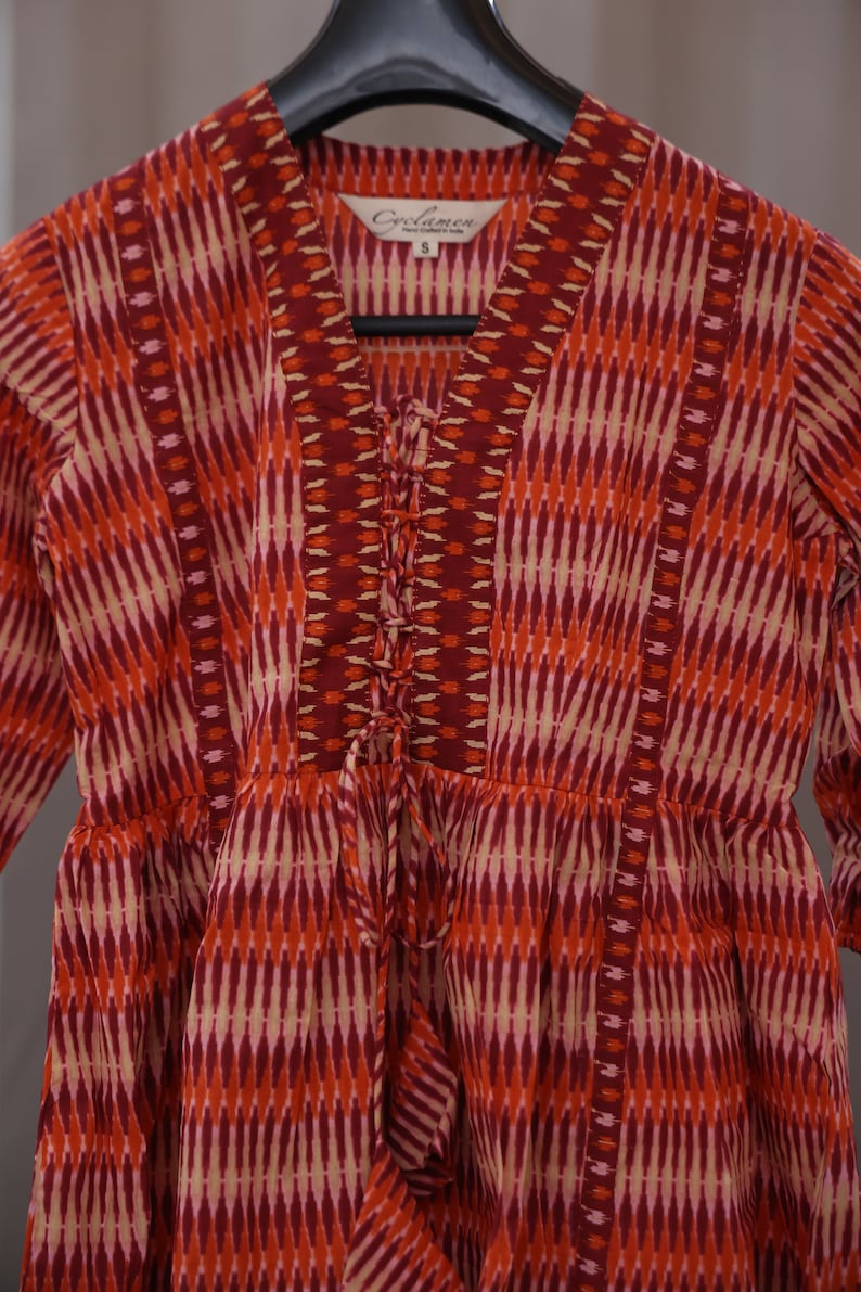 Op de afbeelding: Een rood, oranje en cr&egrave;mekleurige jurk met een vetersluiting aan de voorkant. De jurk heeft een V-hals en is gemaakt van een lichtgewicht stof. Het label op de jurk staat "Coolmen" en "S".