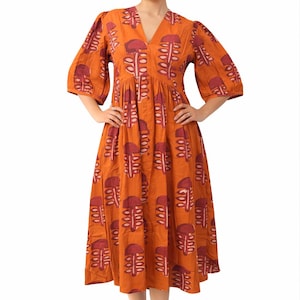 May include: An orange midi dress with a V-neck and elbow-length sleeves. The dress features a repeating pattern of maroon abstract shapes. The model is wearing white sandals. The dress is made of a lightweight fabric.