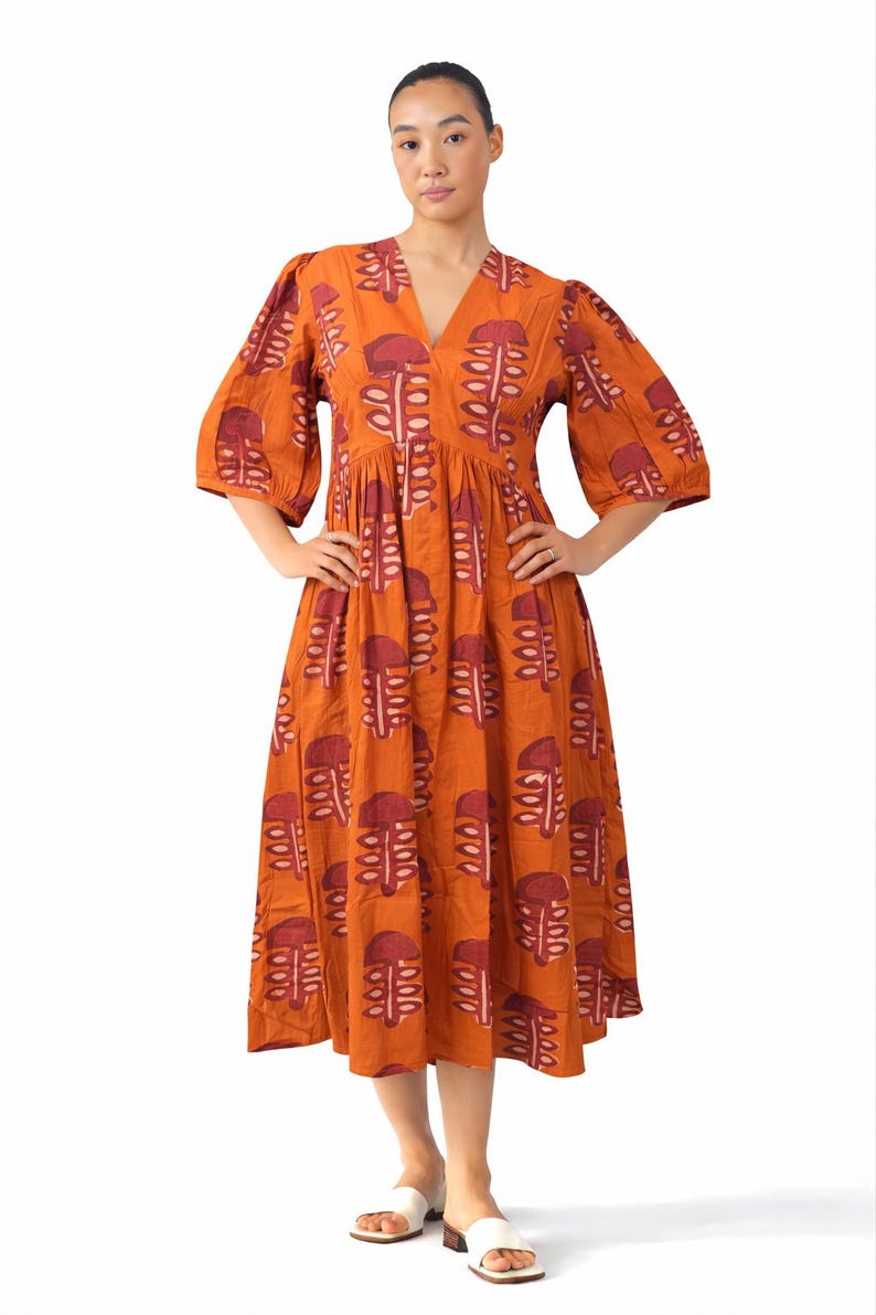 May include: An orange midi dress with a V-neck and elbow-length sleeves. The dress features a repeating pattern of maroon abstract shapes. The model is wearing white sandals. The dress is made of a lightweight fabric.