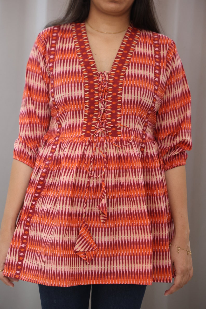 Op de afbeelding: Een rood, oranje en wit gepatroneerde tuniek met een vetersluiting aan de halslijn. De tuniek heeft een losse pasvorm en 3/4 mouwen.