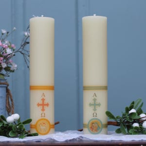 May include: Two ivory-colored pillar candles with decorative bands and religious symbols. One candle has an orange cross and the other has a green cross. Floral arrangements and greenery surround the candles.
