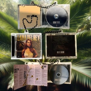 Puede incluir: Carcasas de CD de plástico transparente con cadenas de cuentas negras. Las carcasas muestran el álbum "Born to Die" de Lana Del Rey. La portada del álbum presenta a una mujer con cabello castaño largo. Las carcasas de CD están abiertas.
