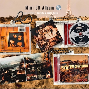 Puede incluir: Mini álbum de CD con la carátula del álbum "Toxicity" de System of a Down. Las fundas de plástico transparente contienen el CD y la carátula. El texto "Mini CD Album" y "Tap-to-Play Spotify & Apple Music" son visibles.