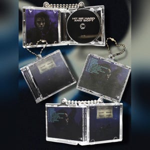 May include: Clear acrylic CD-shaped keychains. One keychain is open, displaying a black CD with the text "HIT ME HARD AND SOFT." Other keychains show album art and track listings. The album art features a person with dark hair.