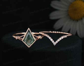 May include: Two rose gold rings. One ring features a green, kite-shaped stone with a delicate design. The second ring is a V-shaped band with small, clear stones. The rings are displayed on a black surface.