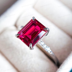 May include: A close-up of a silver ring featuring a large, rectangular, vibrant red gemstone. The ring's band is adorned with small, clear accent stones. The ring is set against a soft, neutral background.
