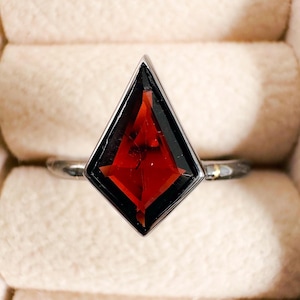 May include: A silver ring featuring a dark red, faceted gemstone set in a diamond shape.